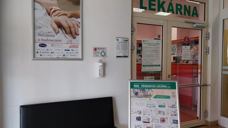 Vstup do lékárny s prosklenými dveřmi, zeleným nápisem „Lékárna“, čekárenskou lavicí a informačním plakátem.